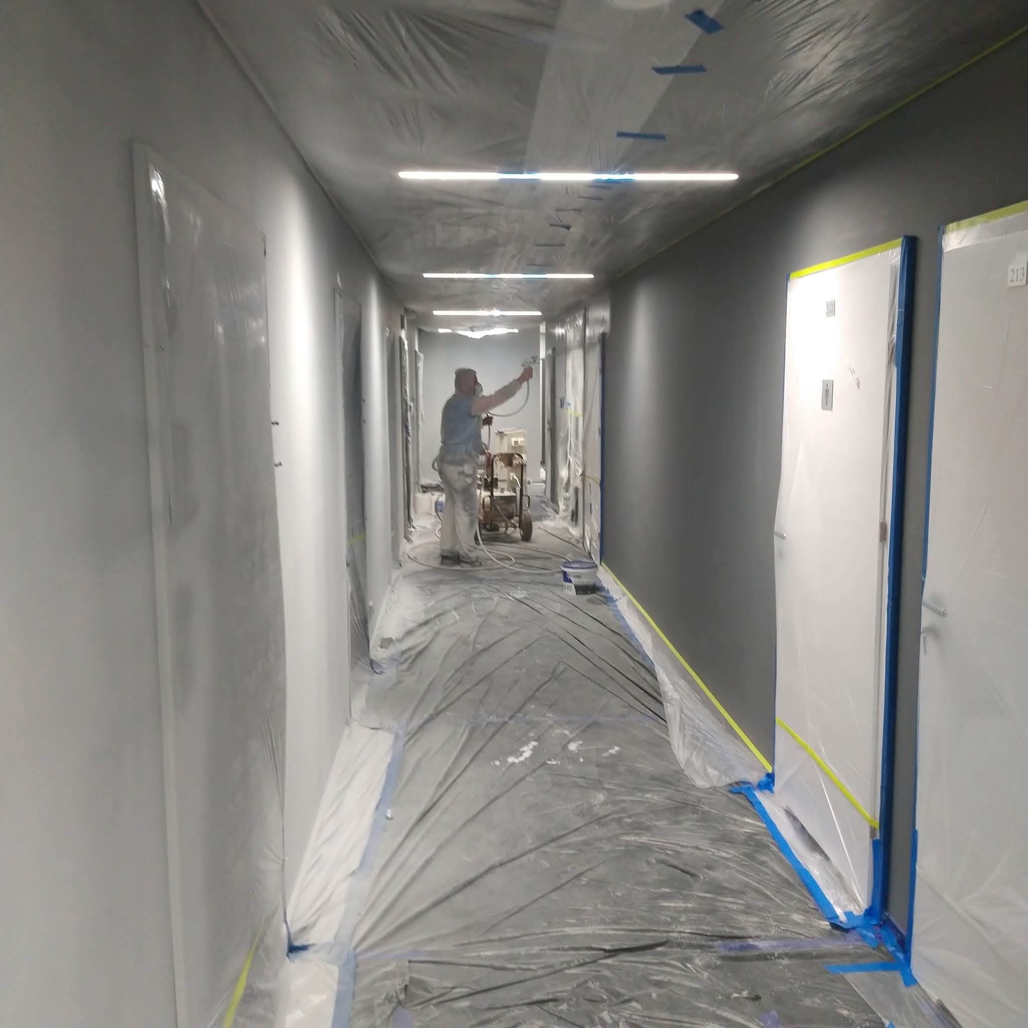 Malowanie natryskowe korytarza biurowego na szaro. Zabezpieczone folią drzwi i podłoga. Pracownik w kombinezonie maluje ściany. Widoczne oświetlenie sufitowe.
