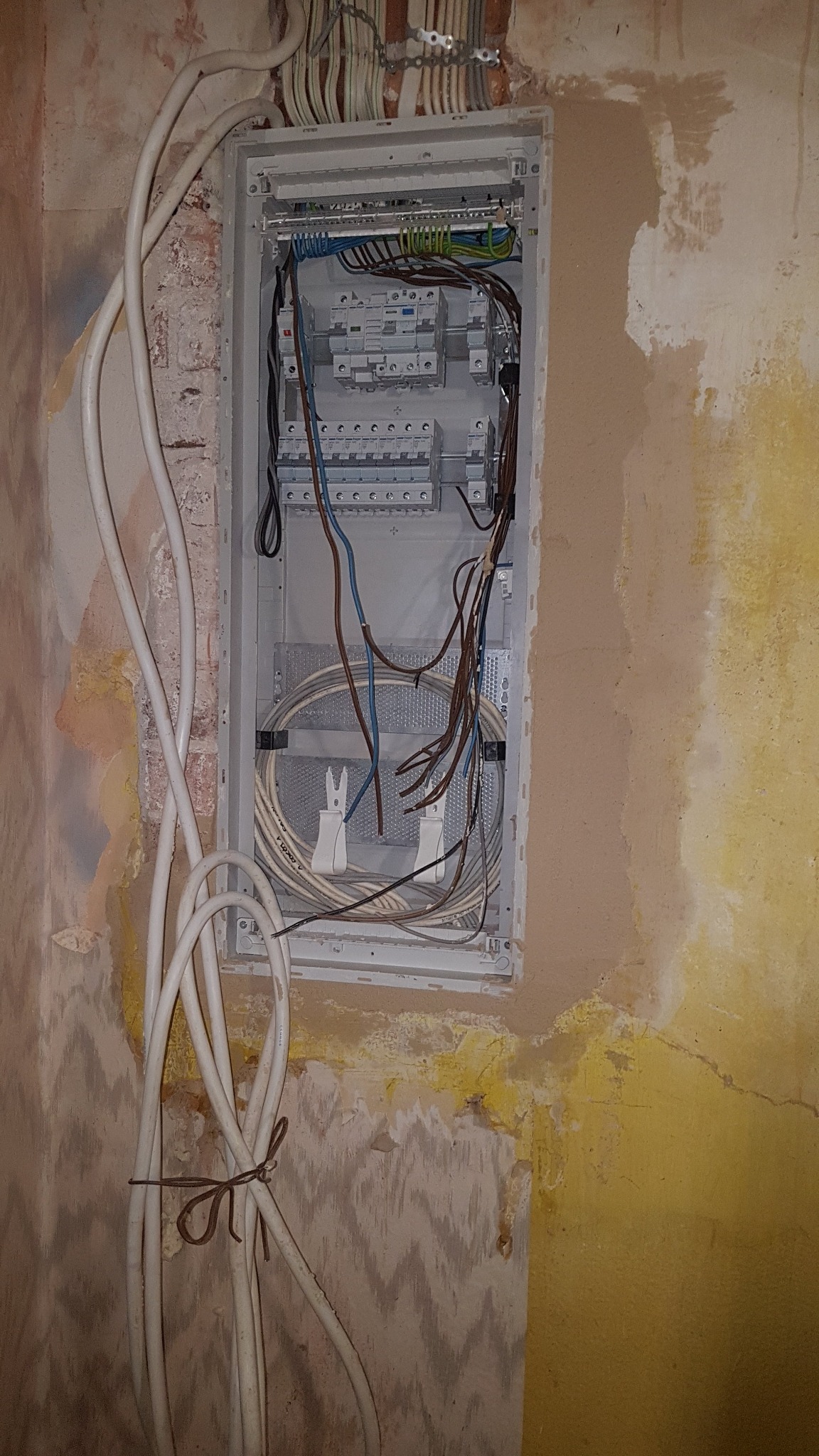 Otwarta skrzynka elektryczna z przewodami na surowej ścianie, widoczne zabezpieczenia i kable wewnątrz oraz wiązka kabli na zewnątrz.