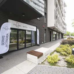 Fasada budynku z szyldem 'PHONE FIX SERWIS AKCESORIA' i dwoma flagami reklamowymi, nowoczesna architektura, zadbana zieleń przed budynkiem.