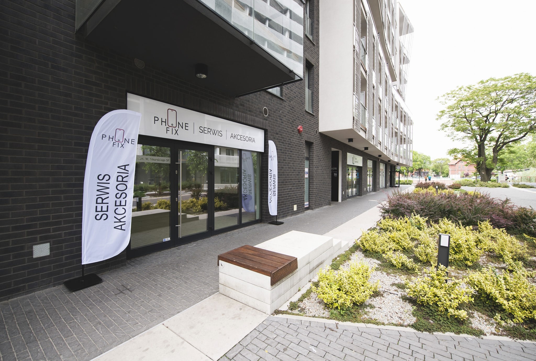 Fasada budynku z szyldem 'PHONE FIX SERWIS AKCESORIA' i dwoma flagami reklamowymi, nowoczesna architektura, zadbana zieleń przed budynkiem.