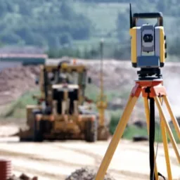 Żółty teodolit na trójnogu ustawiony na placu budowy, w tle pracująca maszyna budowlana i osoba w pomarańczowej kamizelce.