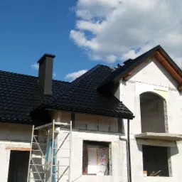 Dom w trakcie budowy z zamontowaną czarną blachą trapezową na dachu, widoczne rusztowanie i fragment rynny, błękitne niebo w tle.