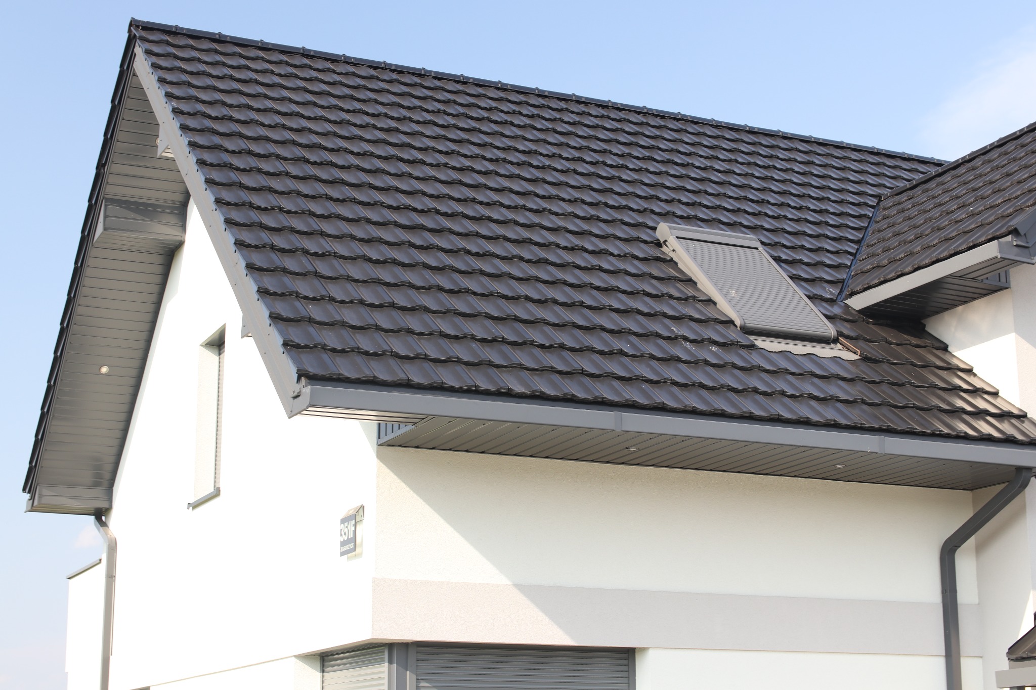 Nowoczesny dom z ciemnoszarą dachówką ceramiczną i oknem dachowym z żaluzjami, widok na fragment elewacji z rynnami i numerem budynku.