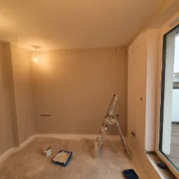 Pokój w trakcie prac: świeżo położone gładzie na ścianach, drabina aluminiowa, kuweta z farbą i wiaderko, widoczne gniazdka elektryczne, okno balkonowe z widokiem na miasto.
