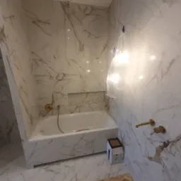Nowa łazienka z wanną obudowaną białymi płytkami imitującymi marmur, złota armatura, karton na podłodze i folia ochronna na ścianie.
