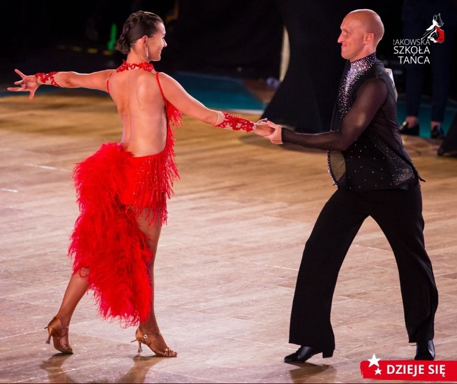 Para tancerzy w strojach konkursowych prezentuje taniec towarzyski na parkiecie, widoczna czerwona suknia z frędzlami i piórami oraz czarny strój tancerza z błyszczącymi aplikacjami.