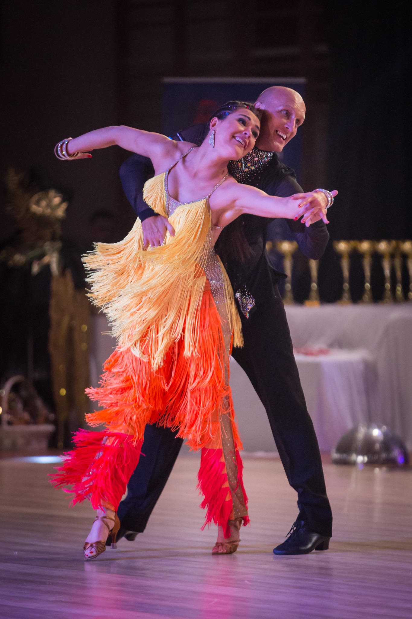 Para tancerzy w dynamicznej pozie, kobieta w sukience z frędzlami ombre, mężczyzna w czarnym stroju, na parkiecie, w tle trofea.