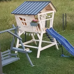 Drewniany domek dla dzieci na podwyższeniu z niebieskim dachem, zjeżdżalnią i huśtawką, udekorowany girlandą z trójkątów i kwiatami w doniczce, na tle zielonego trawnika i nowoczesnego domu.