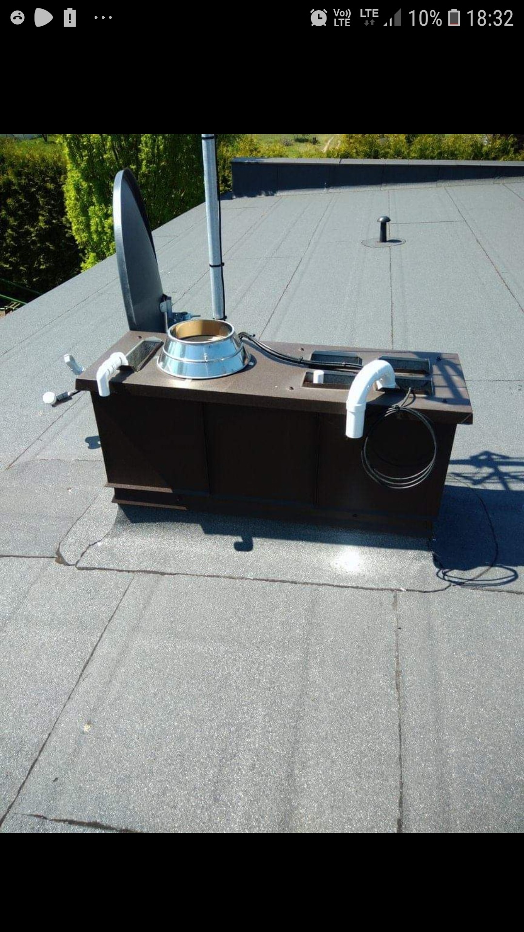 Szary dach pokryty papą z brązową obudową kominka wentylacyjnego, widoczne elementy instalacji i antena satelitarna.