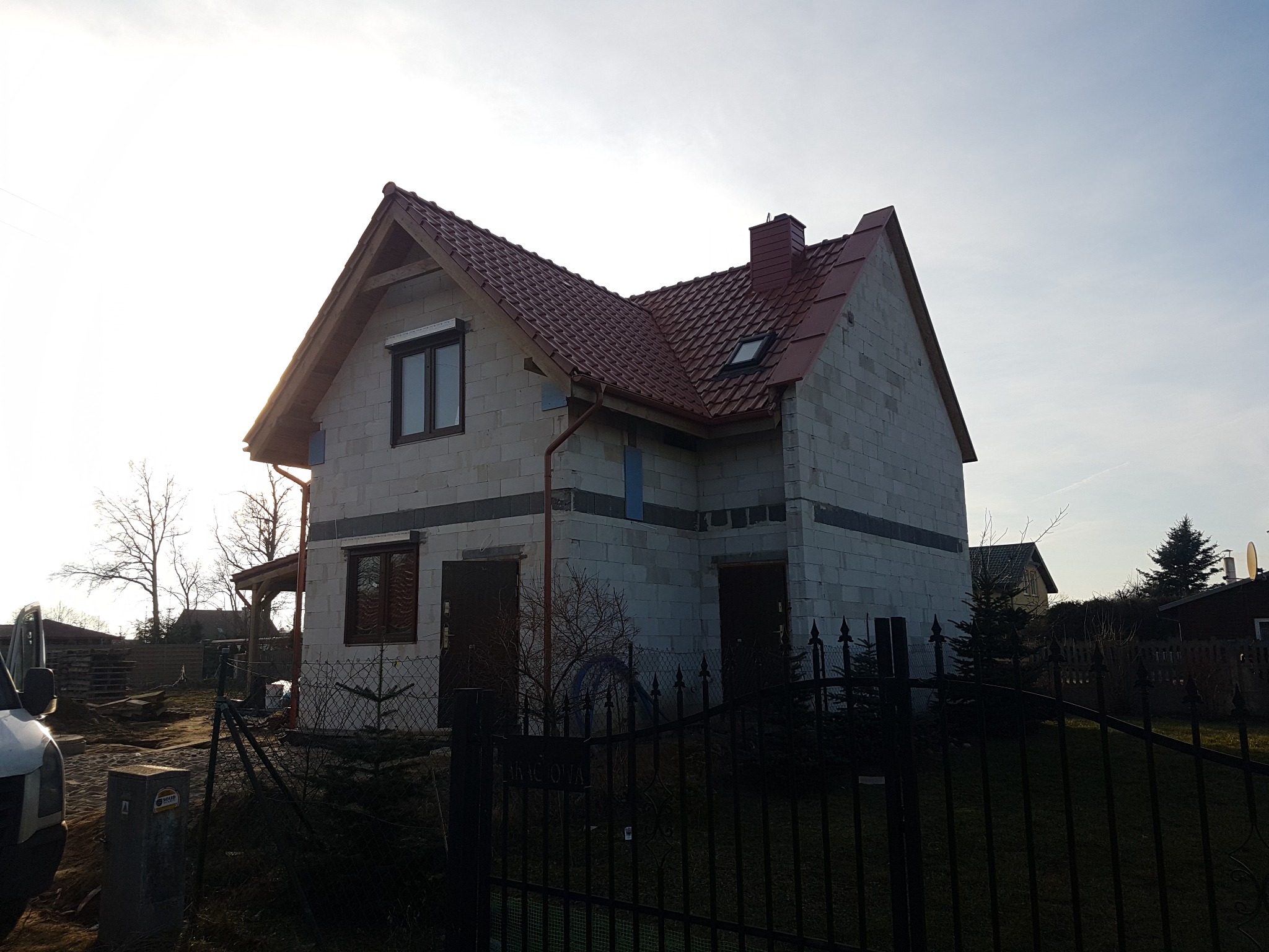Surowy dom jednorodzinny z czerwonym dachem, w trakcie budowy, widok zza metalowego ogrodzenia, widoczne okna z roletami, rynny i komin.
