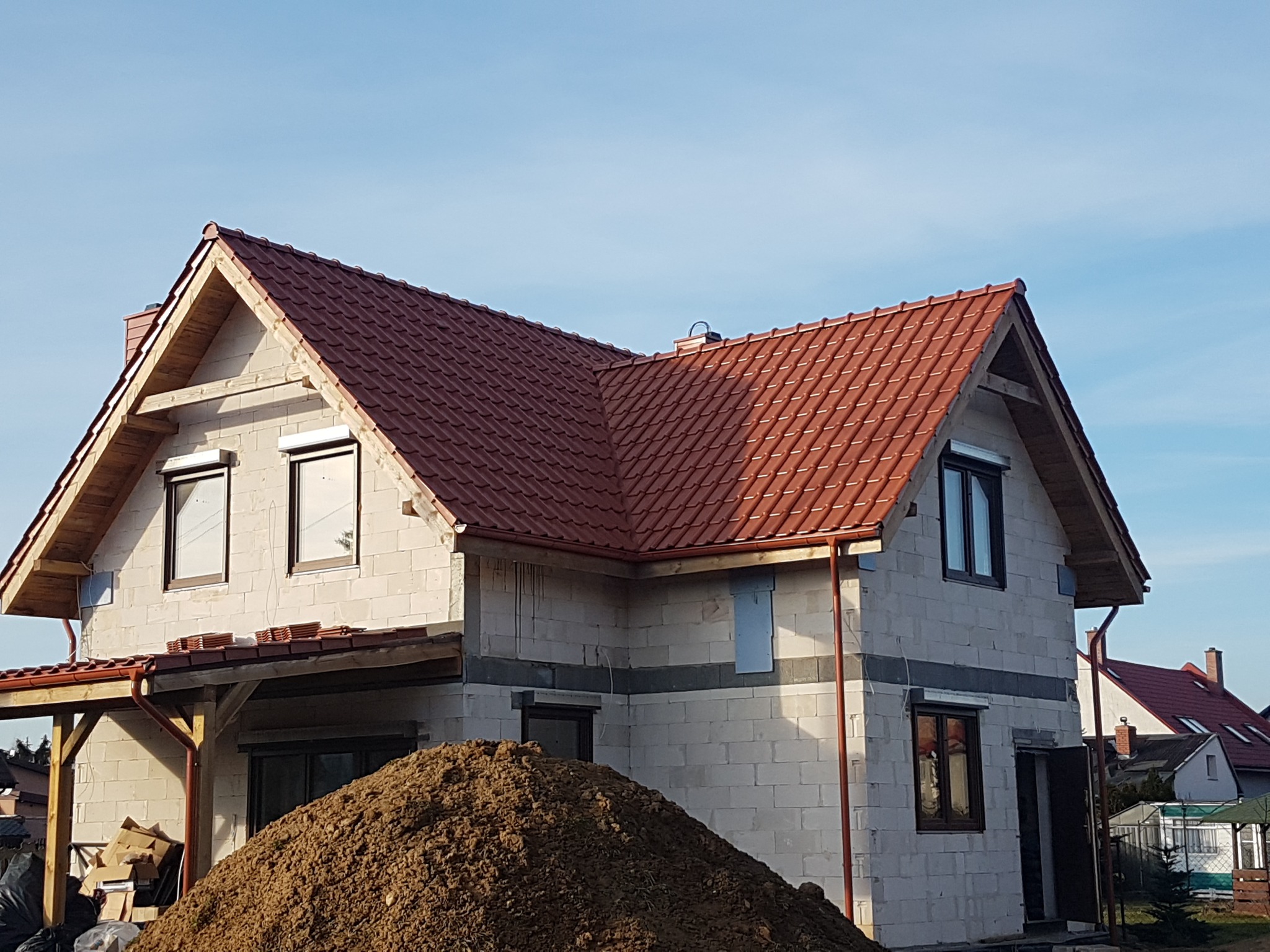 Surowy dom z czerwonym dachem z dachówki ceramicznej w trakcie budowy, widoczna sterta ziemi przed budynkiem, okna z roletami zewnętrznymi.
