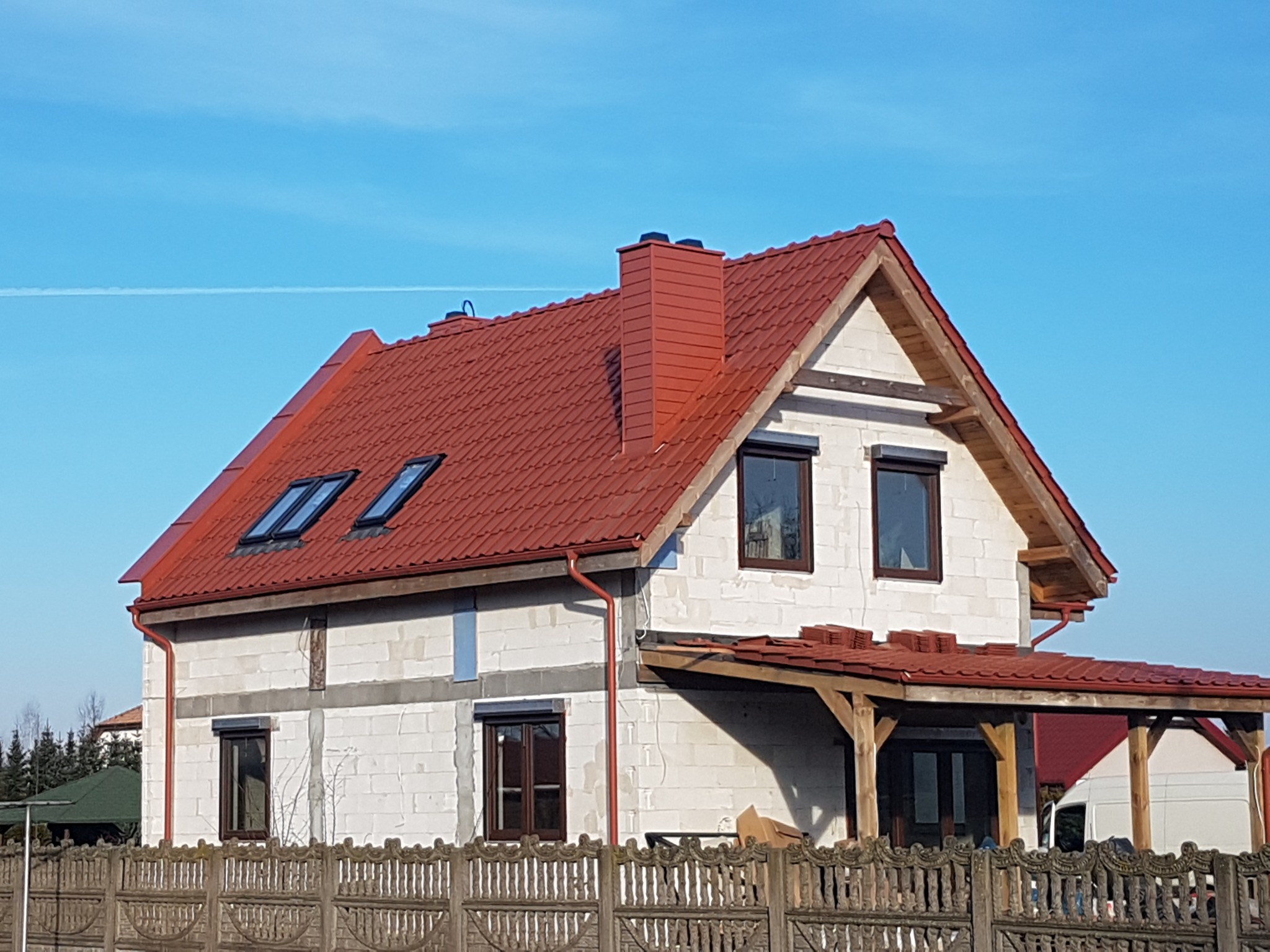 Dom w trakcie budowy z czerwoną dachówką, widoczne okna dachowe, ściany z białego pustaka i drewniana konstrukcja zadaszenia tarasu, ogrodzenie z betonowych przęseł w pierwszym planie, błękitne...
