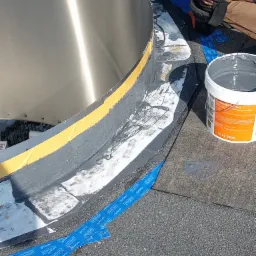 Uszczelnianie obr&oacute;bki blacharskiej komina na dachu płaskim za pomocą płynnej membrany poliuretanowej, widoczny fragment dachu pokrytego papą oraz niebieska taśma zabezpieczająca.