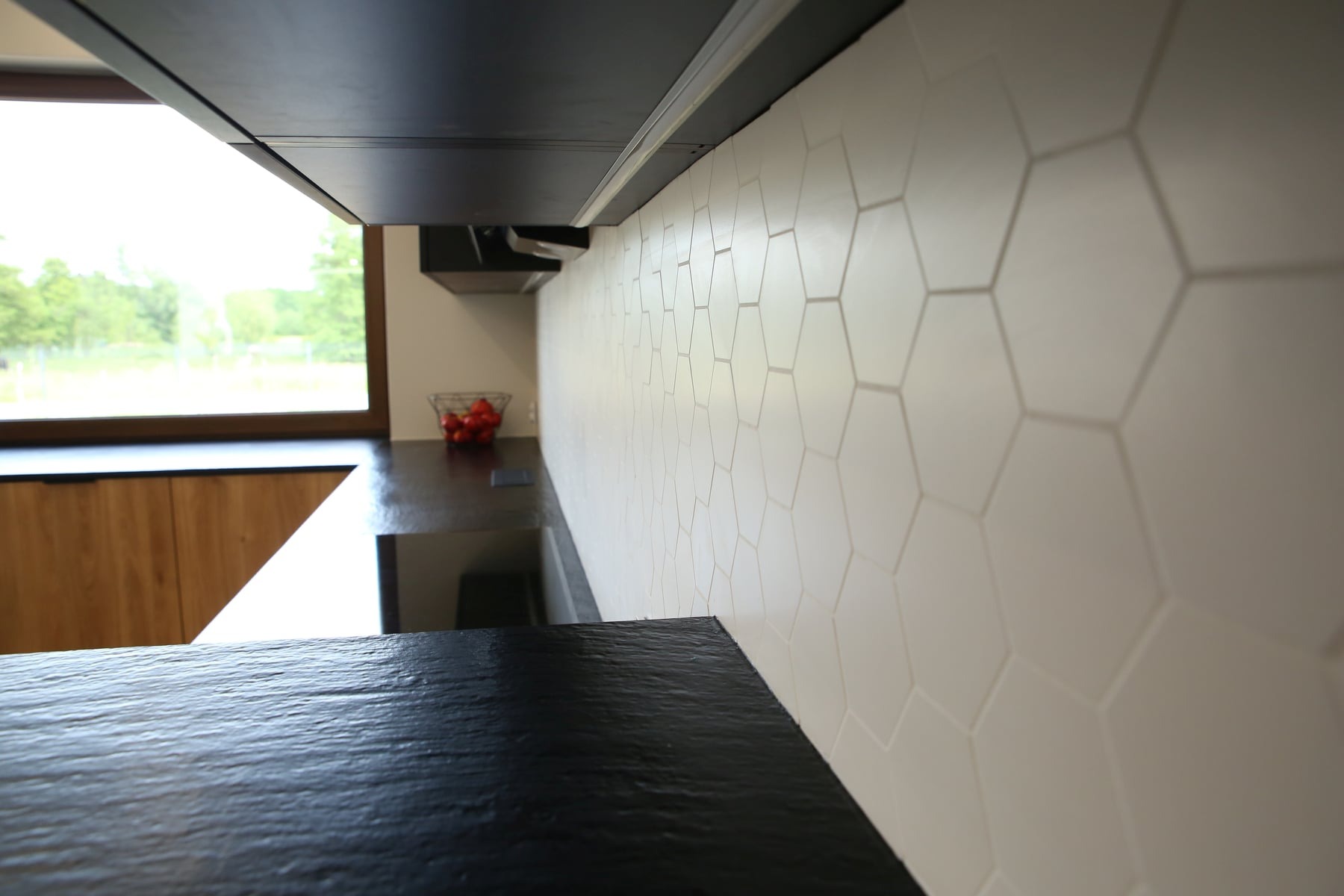 Nowoczesna kuchnia z czarnym blatem, heksagonalnymi płytkami na ścianie i widokiem na zieleń za oknem.