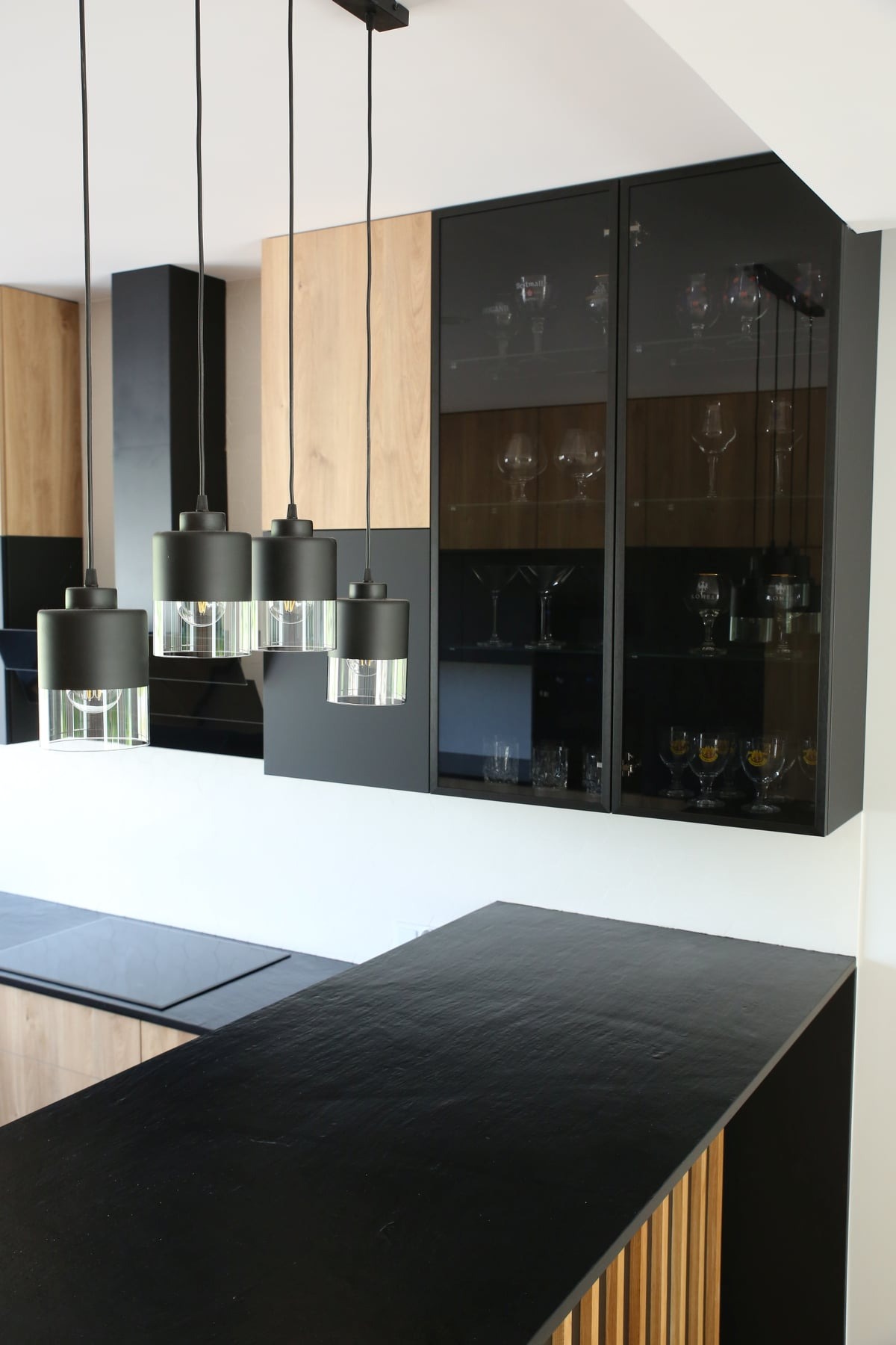 Nowoczesna kuchnia z czarnym blatem z kamienia, wiszącymi lampami z przezroczystymi elementami i szklaną witryną z kieliszkami.