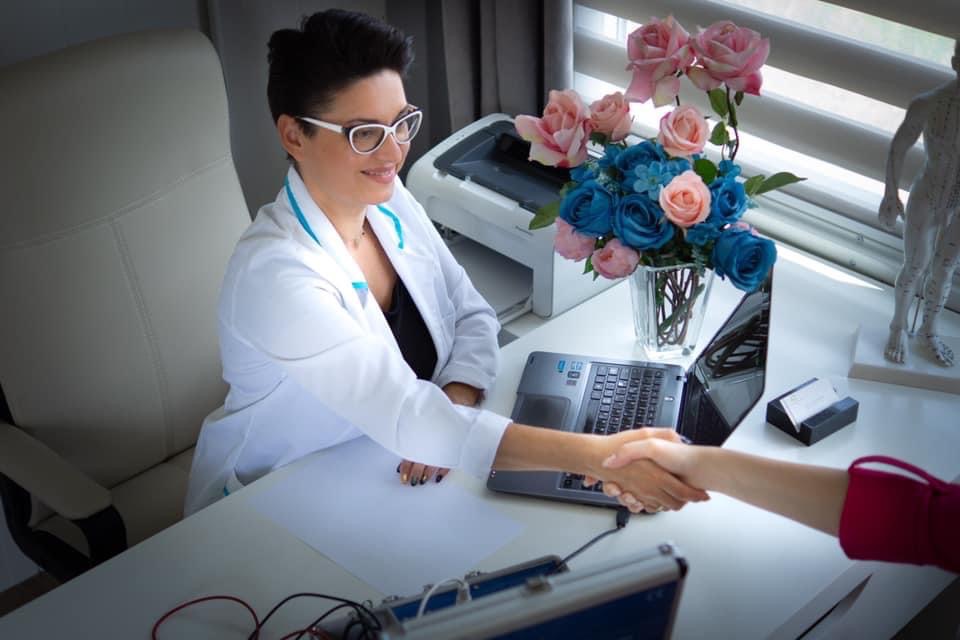 Uśmiechnięta kobieta w białym fartuchu i okularach wita się z pacjentem na jasnym biurku z laptopem, kwiatami w wazonie i modelem akupunkturowym w tle.