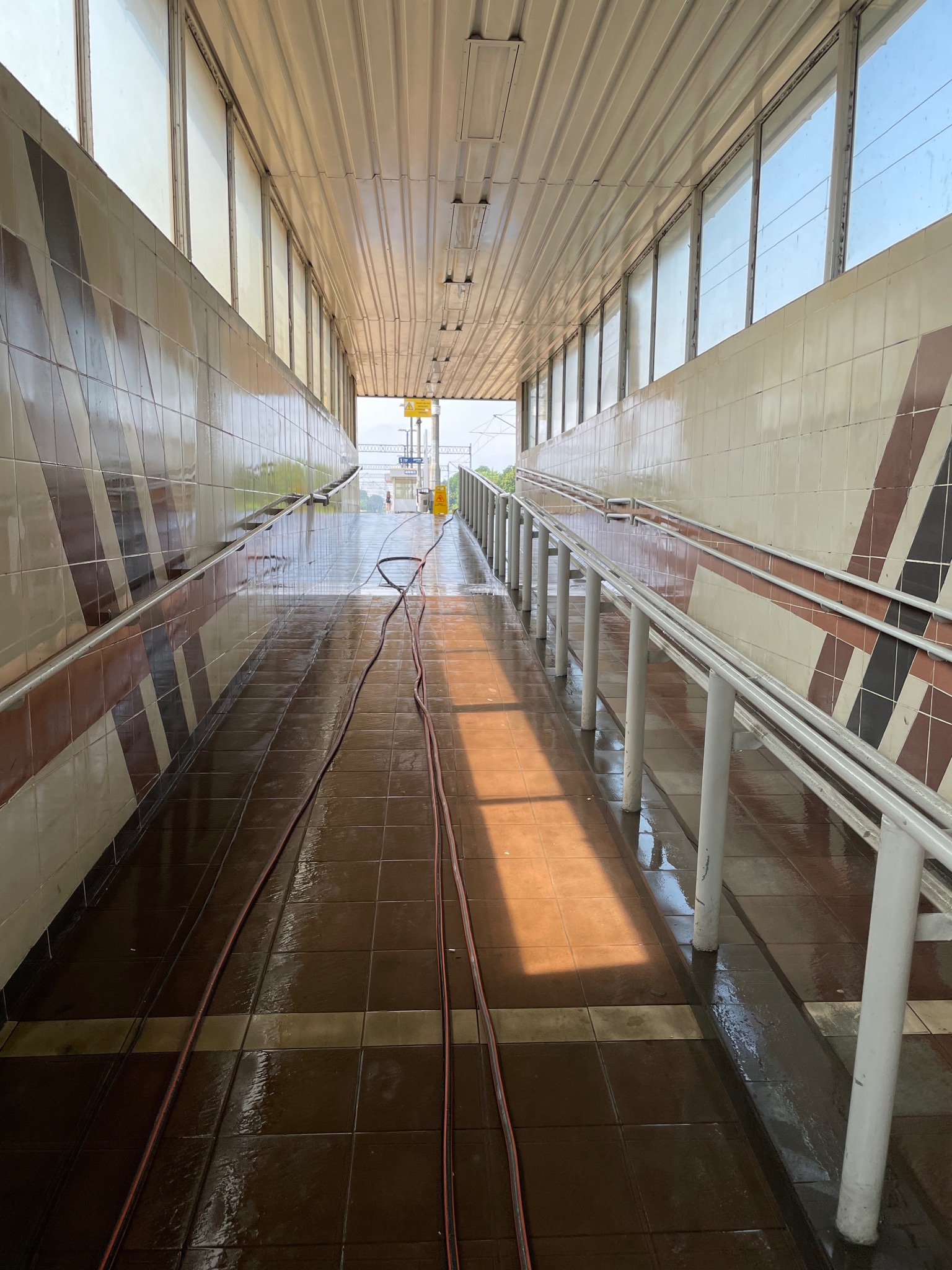 Mokry, kafelkowany korytarz z rampą i metalowymi poręczami, w trakcie mycia pod ciśnieniem, z widocznym wężem ciśnieniowym na podłodze.