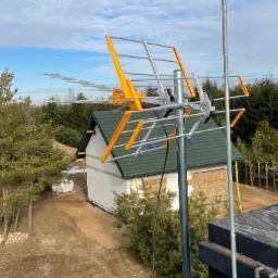 Antena telewizyjna Yagi-Uda z pomarańczowymi elementami na tle budowanego domu z zielonym dachem i lasem iglastym, widoczna z dachu budynku.