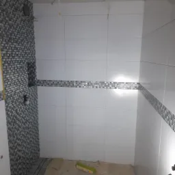 Wnętrze łazienki w trakcie remontu: białe płytki ścienne ułożone do pewnej wysokości, z dekoracyjnym pasem mozaiki, na podłodze widoczne resztki zaprawy i szczotka.