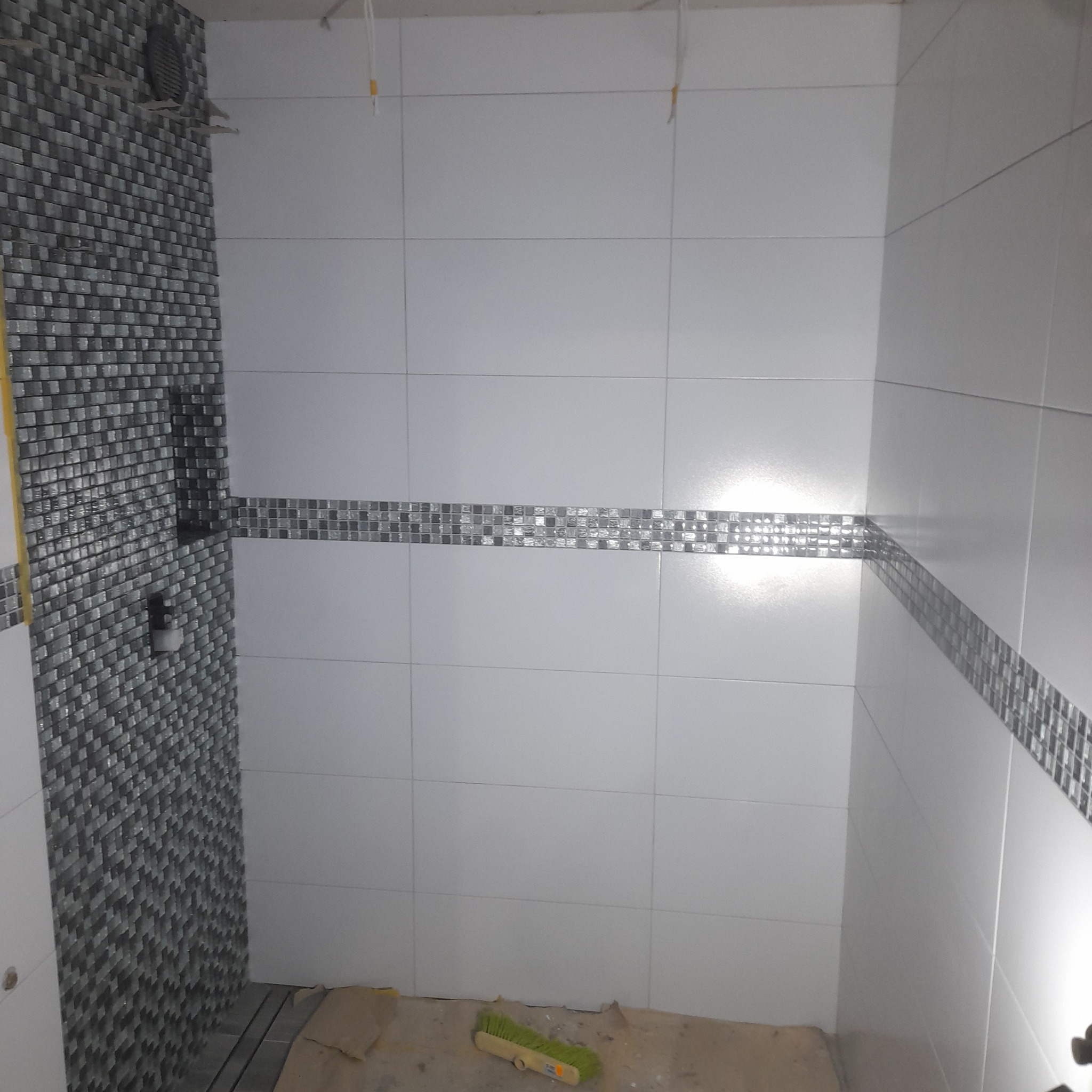 Wnętrze łazienki w trakcie remontu: białe płytki ścienne ułożone do pewnej wysokości, z dekoracyjnym pasem mozaiki, na podłodze widoczne resztki zaprawy i szczotka.