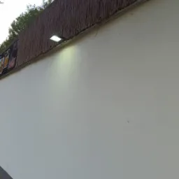 Zewnętrzny reflektor LED oświetla białą ścianę z dekoracyjnym, trzcinowym panelem na górze. Widoczna kostka brukowa u dołu i fragment nieba.