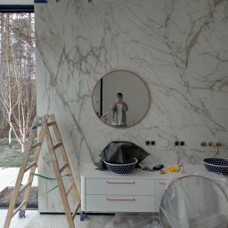 FRBIW Damian Lumer - Łazienka w trakcie wykańczania: marmurowe płytki, okrągłe lustro z miedzianą ramą, biała szafka z miedzianymi uchwytami, drabina malarska i akcesoria na blacie.