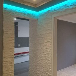 FRBIW Damian Lumer - Korytarz z dekoracyjną ścianą z kamienia, podświetlony niebieskim światłem LED, widoczne drzwi do innego pomieszczenia i fragment podłogi z płytek.