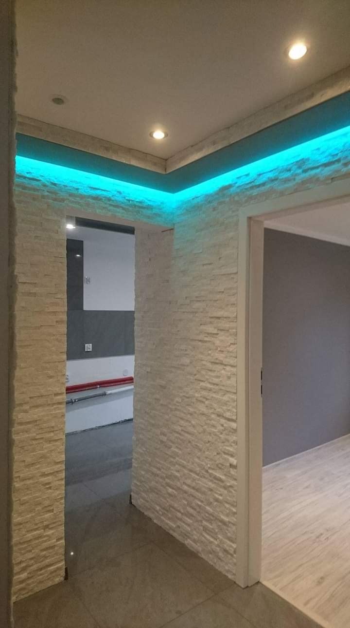 Korytarz z dekoracyjną ścianą z kamienia, podświetlony niebieskim światłem LED, widoczne drzwi do innego pomieszczenia i fragment podłogi z płytek.