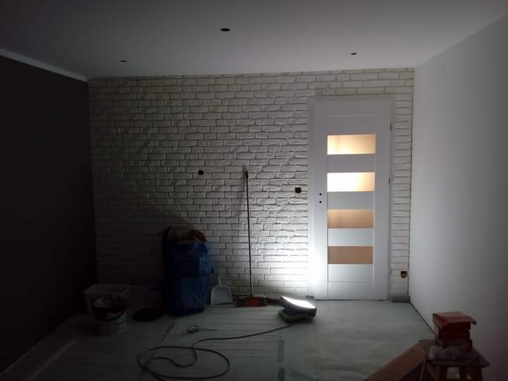 Pokój w trakcie wykańczania: ściana z białej cegły dekoracyjnej, białe drzwi z przeszkleniami, ciemna ściana obok, widoczne narzędzia i akcesoria malarskie, tymczasowe oświetlenie podłogowe.