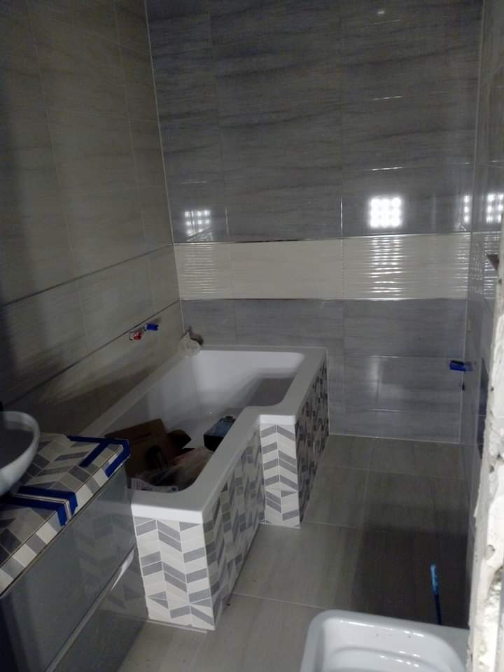 Wnętrze łazienki w trakcie remontu, widoczna wanna z obudową w szary wzór chevron, szare płytki na ścianach i podłodze, oraz niedokończone elementy wykończeniowe.