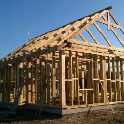 Surowy, drewniany szkielet domu w trakcie budowy, z widoczną konstrukcją dachu i ścian, na tle błękitnego nieba i zalesionego krajobrazu.