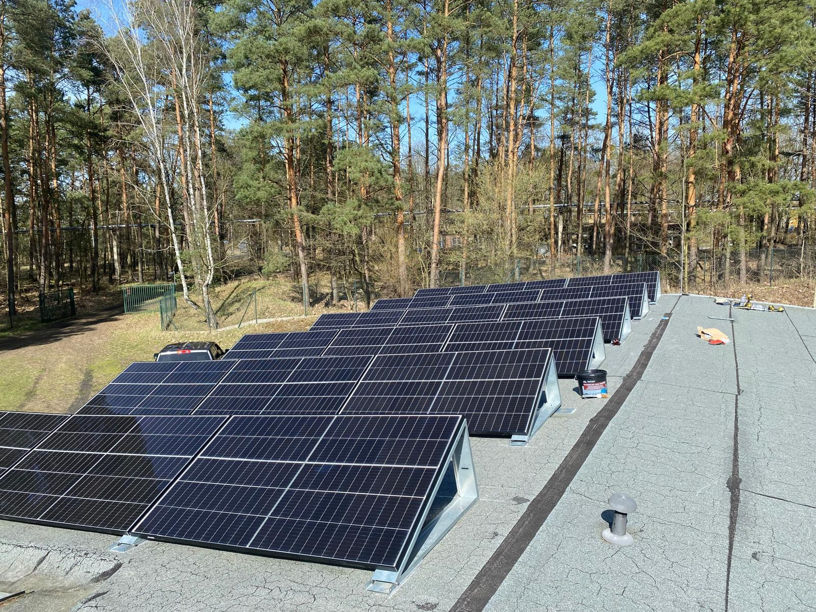 Rząd paneli fotowoltaicznych na płaskim dachu pokrytym papą, z lasem w tle. Widoczne narzędzia i puszka z uszczelniaczem. Instalacja w trakcie montażu.