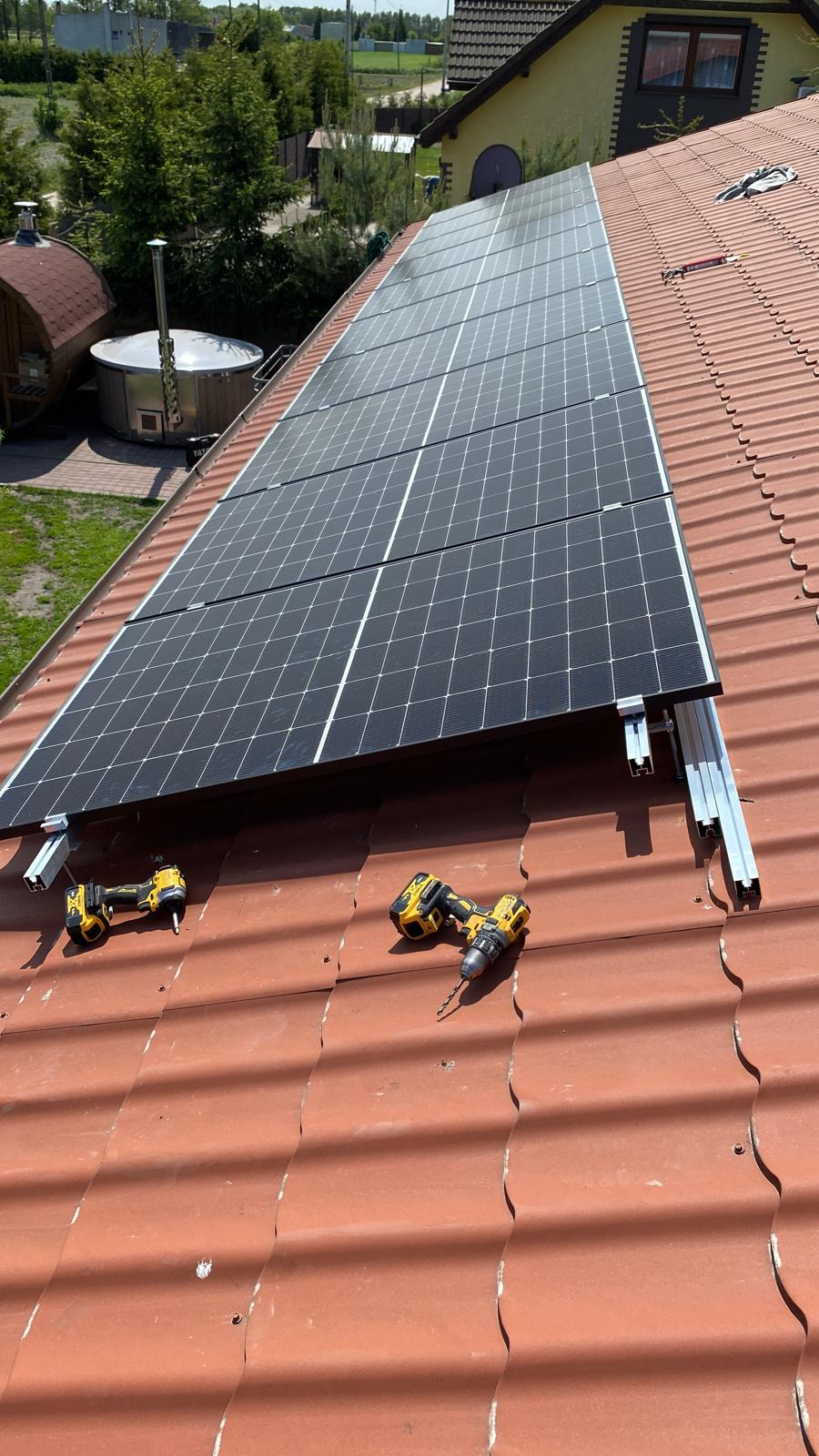 Montaż paneli fotowoltaicznych na dachu z czerwonej dachówki. Widoczne dwie wkrętarki akumulatorowe, szyny montażowe i częściowo zamontowane panele.