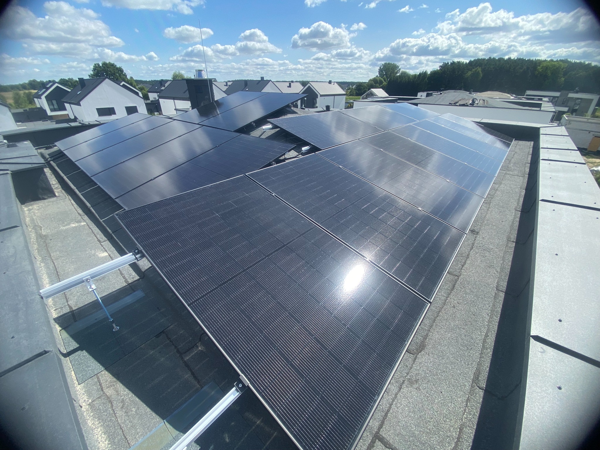 Błyszczące panele fotowoltaiczne na dachu budynku mieszkalnego w słoneczny dzień. Widoczne mocowania paneli i fragmenty dachu. W tle zabudowa jednorodzinna i błękitne niebo.