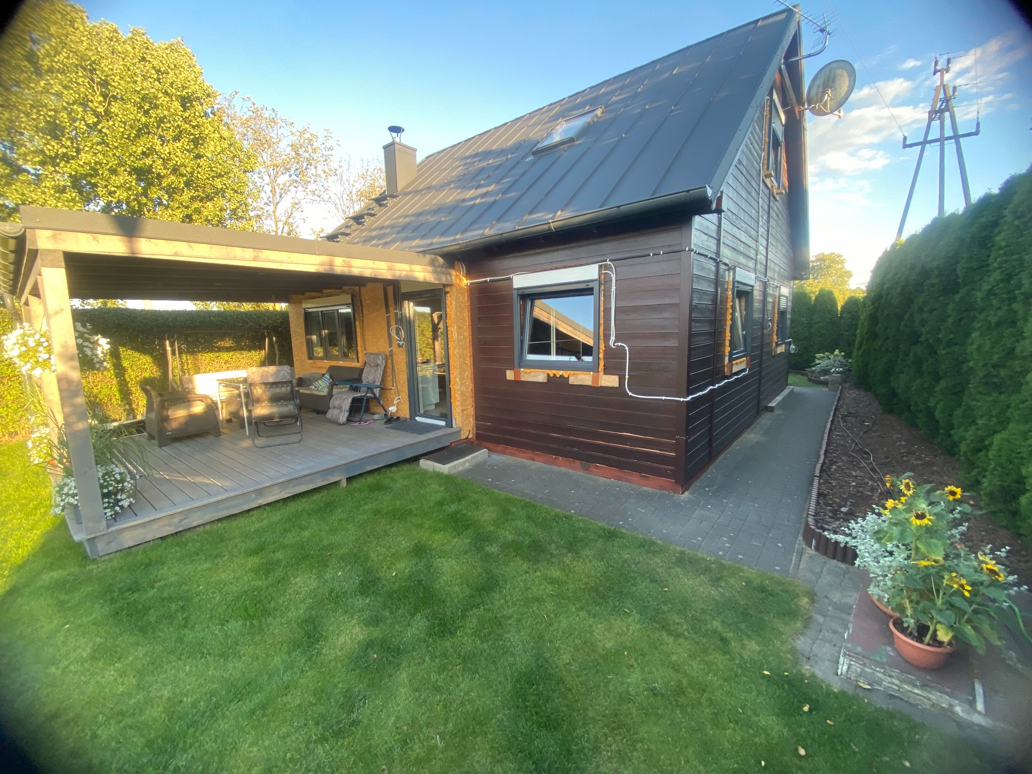 Brązowy dom szkieletowy z ciemnym dachem i zadaszonym tarasem, otoczony zielonym trawnikiem i roślinnością. Widoczne okna z roletami zewnętrznymi i instalacje na elewacji.