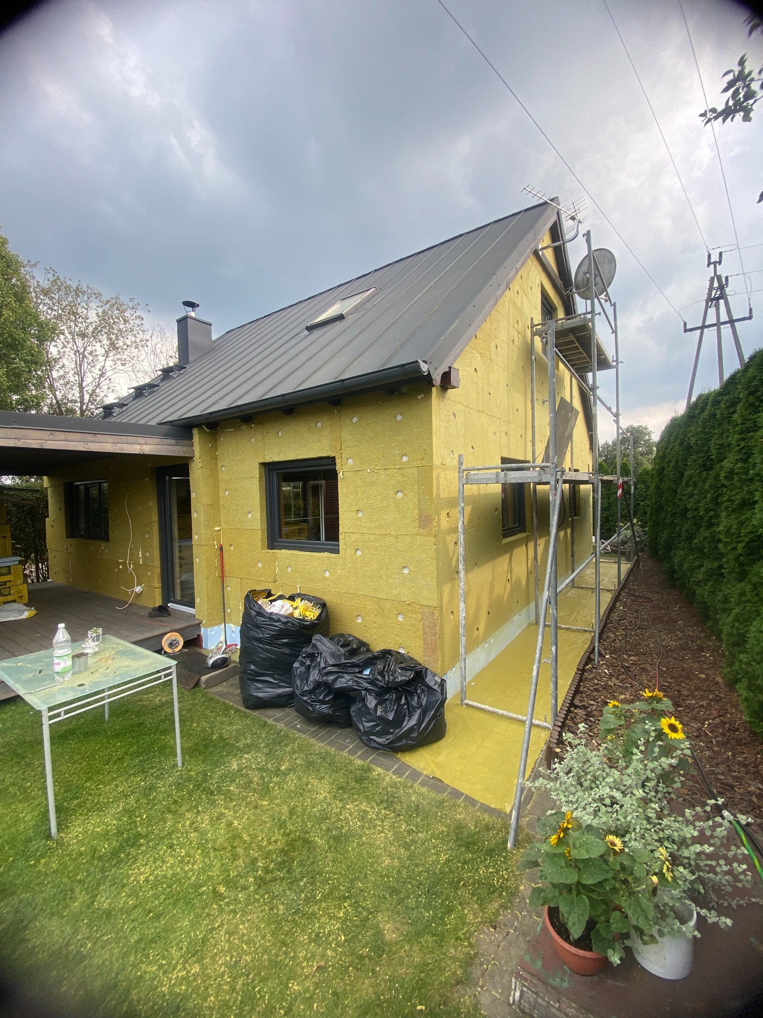 Ocieplanie domu wełną mineralną, widoczne rusztowania i worki z odpadami. Dom z szarym dachem, okna w czarnych ramach. Prace w toku, widok z ogrodu.