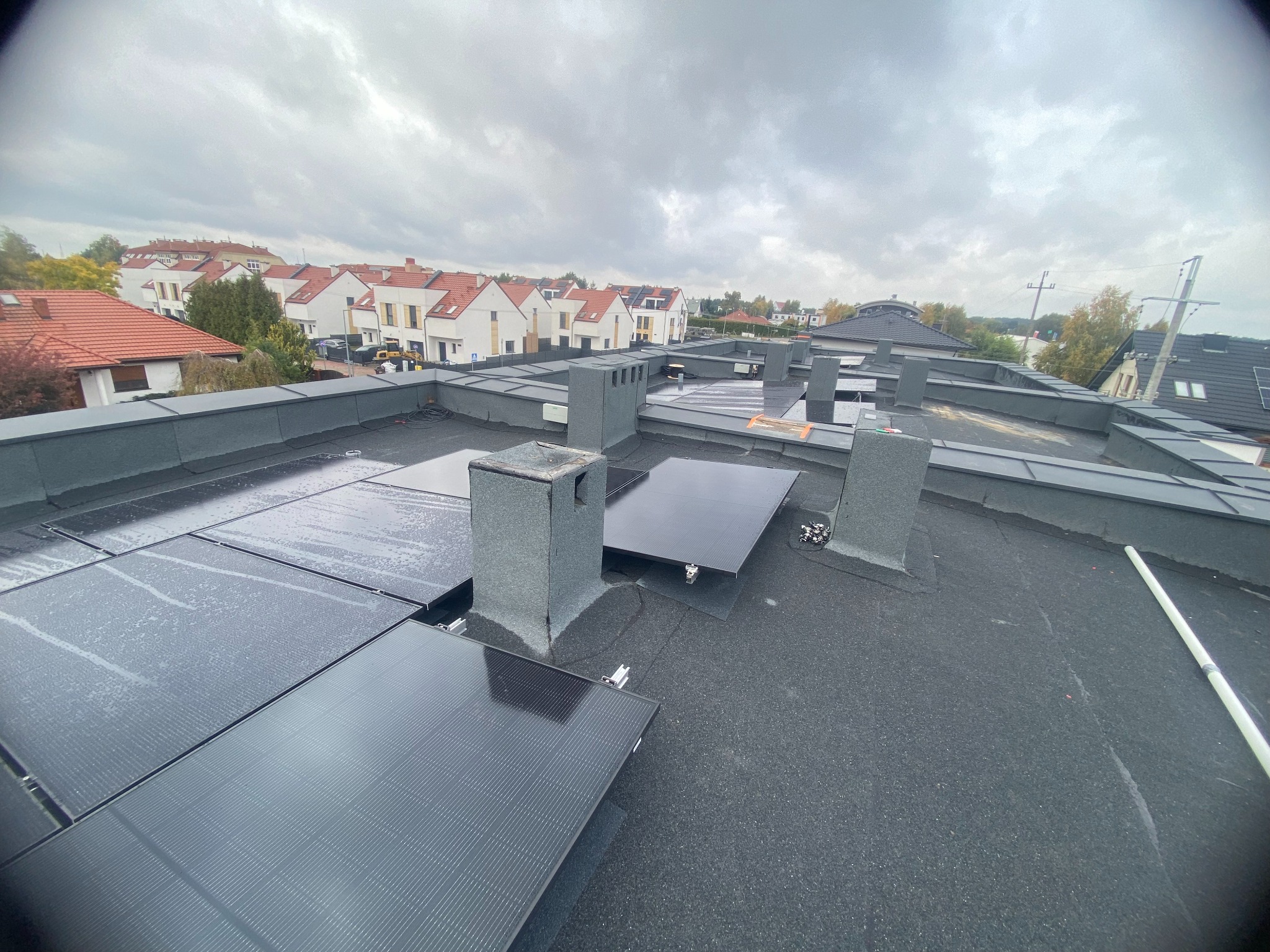 Montaż paneli fotowoltaicznych na dachu z kominami, widok z góry na osiedle domów jednorodzinnych w pochmurny dzień. Widoczne detale mocowań paneli.