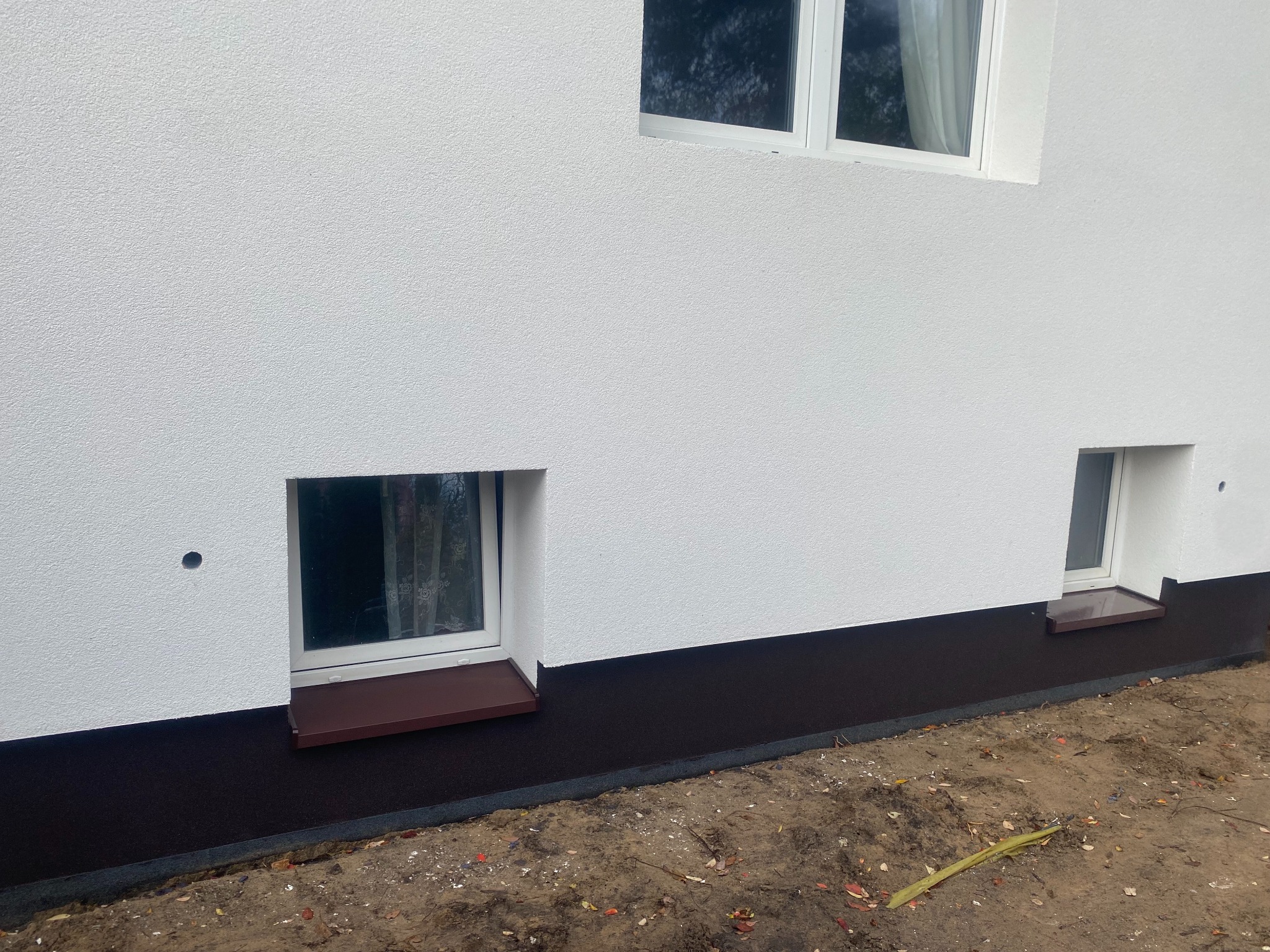 Biała elewacja budynku z trzema oknami, z brązowym pasem cokołu. Widoczne okna piwniczne z brązowymi parapetami. Tynk strukturalny.