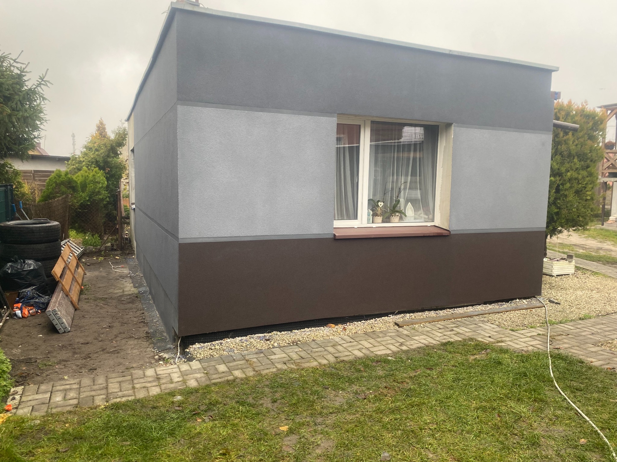 Świeżo otynkowany budynek gospodarczy z oknem, pomalowany w trzech odcieniach szarości i brązu, otoczony zielenią i kostką brukową. Widoczne narzędzia i materiały budowlane.