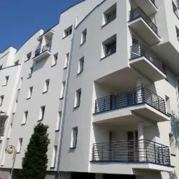 Biały budynek wielorodzinny z balkonami i rusztowaniami, w słoneczny dzień. Nowoczesna architektura z wyraźnymi, geometrycznymi liniami.