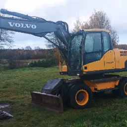 Żółto-szara koparka Volvo EW140C na kołach, stojąca na trawiastym terenie z widocznymi drzewami i pochmurnym niebem w tle, z datą 23.10.2021 w prawym dolnym rogu.