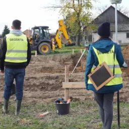 Dwóch pracowników w kamizelkach odblaskowych z napisem, podczas prac ziemnych na budowie, w tle koparka JCB i budynek gospodarczy.