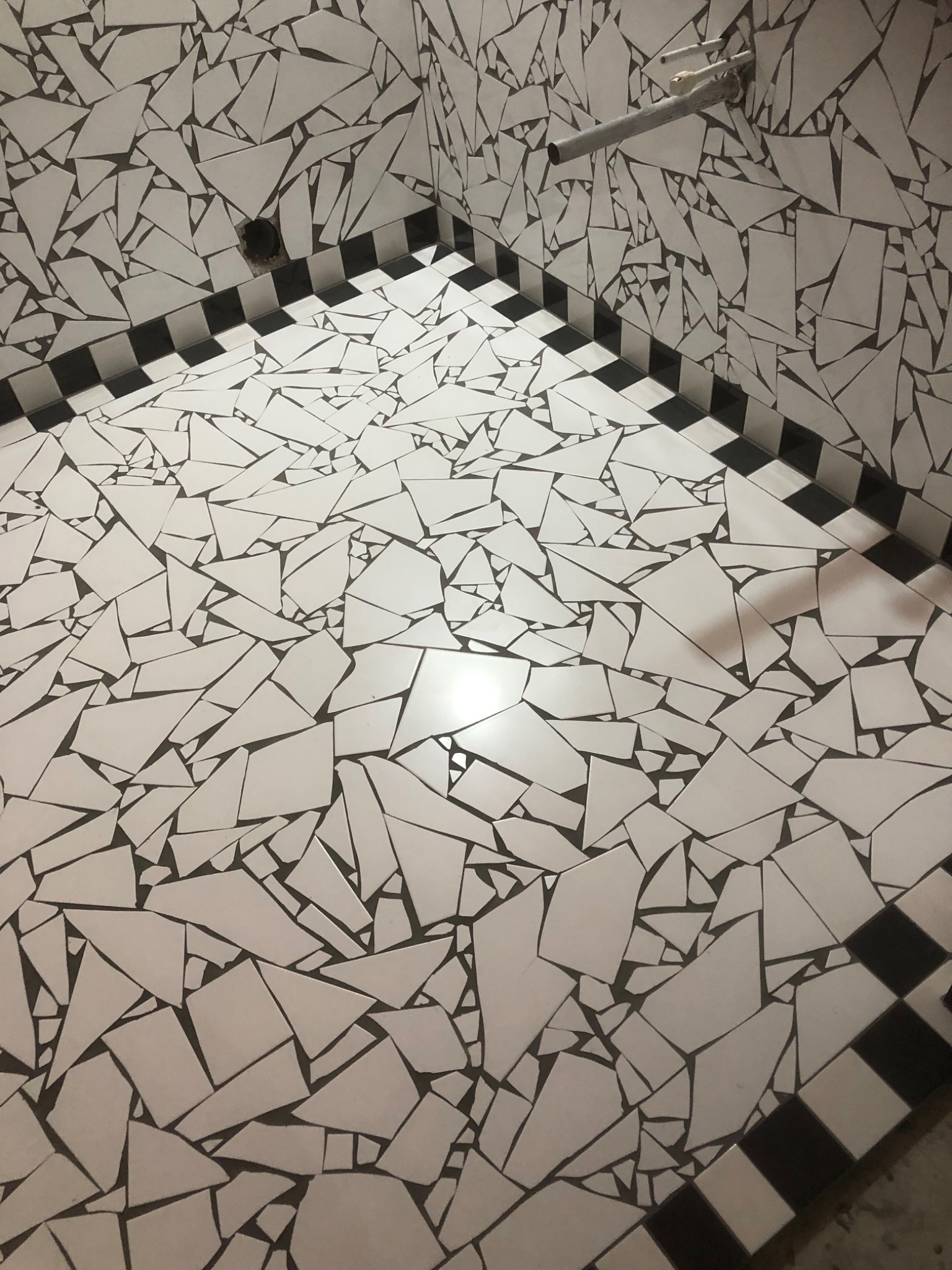 Ściana i podłoga w łazience wyłożone nieregularnymi, białymi płytkami z ciemnymi fugami, z czarno-białym pasem płytek na styku ściany i podłogi.