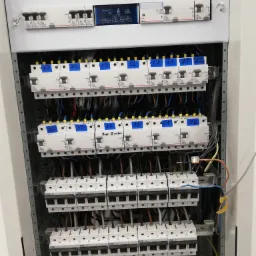 Rozdzielnica elektryczna z zamontowanymi bezpiecznikami, oznaczonymi numerami na niebieskich naklejkach, widoczne przewody i złączki, elementy instalacji w trakcie montażu.