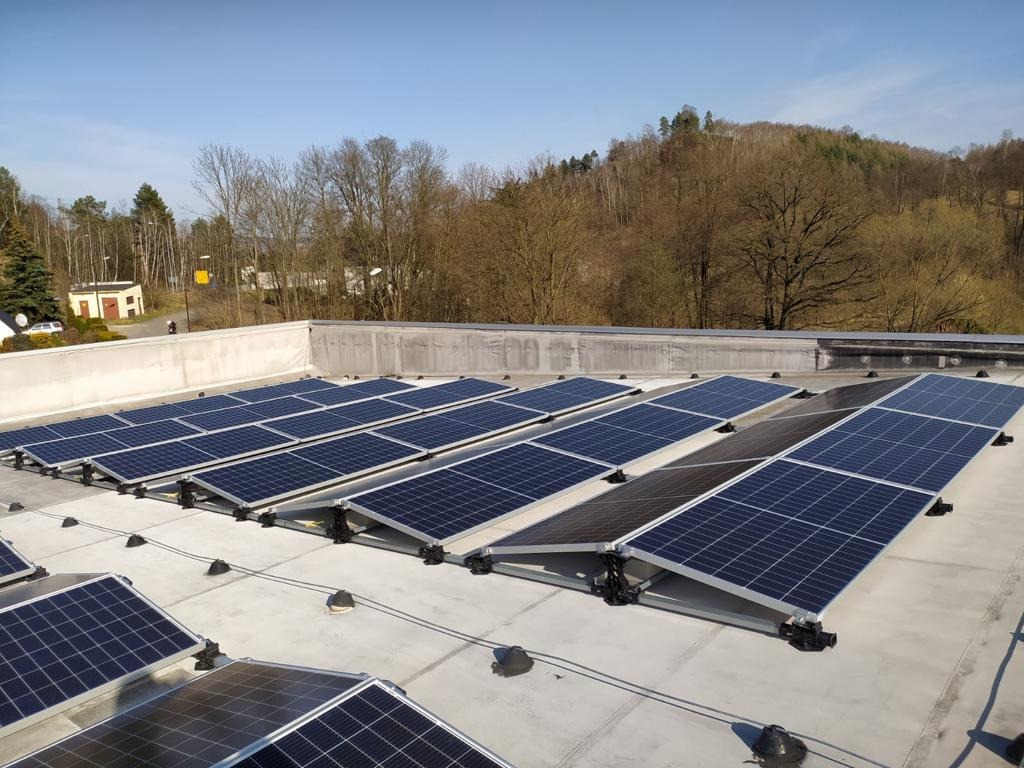 Rząd paneli słonecznych na płaskim dachu budynku, widok z góry, w tle drzewa i pagórki, błękitne niebo.