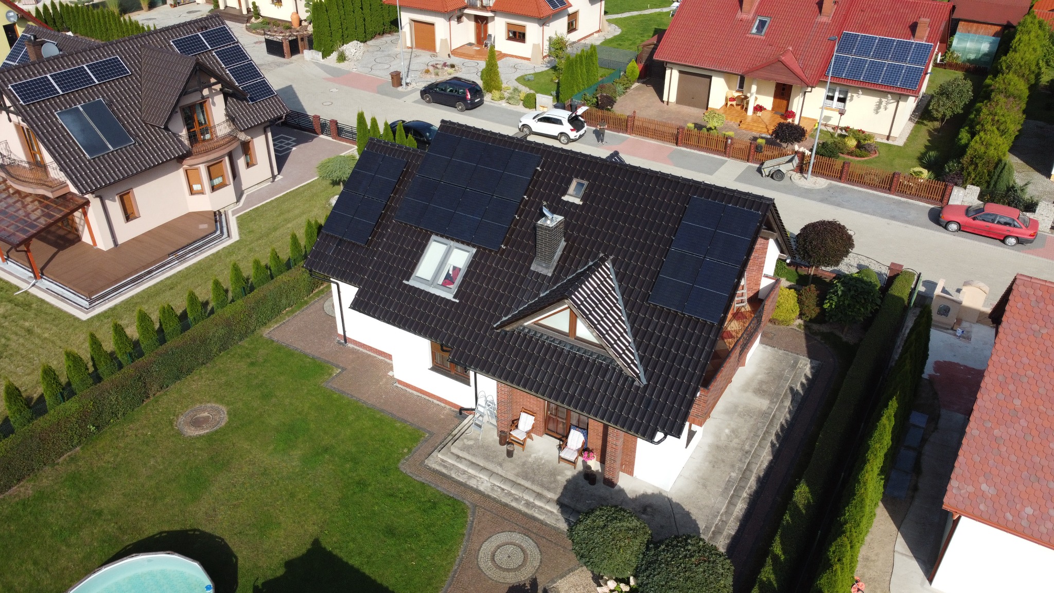 Widok z góry na dom jednorodzinny z ciemną dachówką, na której zamontowane są panele fotowoltaiczne; na dachu widoczny komin i okno dachowe; w tle inne domy i zadbana zieleń.