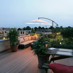 Deski tarasowe Lignodur Terrafina na przestronnym tarasie na dachu w Białych Błotach, z widokiem na miasto o zmierzchu, uzupełnione roślinami w donicach, parasolem i meblami ogrodowymi.