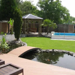 Ogród z oczkiem wodnym, drewnianym mostkiem, tarasem z desek kompozytowych, altaną i basenem, otoczony zielenią i kamieniami dekoracyjnymi.