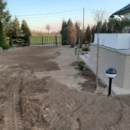 Przygotowany teren pod trawnik z widocznymi śladami opon i butów, w tle ogrodzenie posesji i drzewa iglaste, oświetlenie ogrodowe wzdłuż ścieżki.