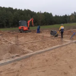 Wykop pod fundamenty z widocznym szalunkiem, wystającymi prętami zbrojeniowymi i zagęszczarką wibracyjną w trakcie pracy oraz minikoparką w tle.